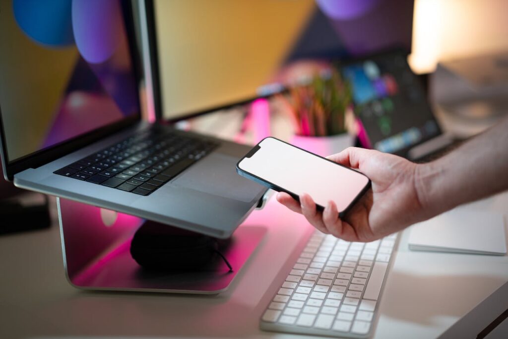 pexels-photo-27861839 Close-up of a hand holding a smartphone with blank screen next to a laptop in a modern office.